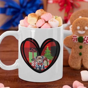 Puede incluir: Taza de cerámica blanca con un diseño en forma de corazón con un cachorro, un árbol de Navidad y regalos. El corazón tiene un patrón de cuadros rojos y negros. La taza está llena de malvaviscos. Una galleta de hombre de jengibre está cerca.