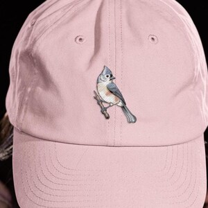 Puede incluir: Una gorra de béisbol de color rosa claro con un diseño bordado de un pájaro carbonero copetudo posado en una pequeña rama marrón. El ave tiene plumaje gris, blanco y rojizo. La gorra tiene una corona estructurada y una visera curva.
