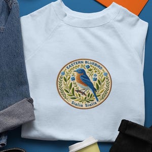 May include: Light blue sweatshirt featuring a circular embroidered patch with an Eastern Bluebird design. The patch includes the text "Eastern Bluebird" and "Sialia Sialis", surrounded by floral and leaf elements. The bird is blue and orange.