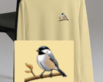 May include: A long-sleeved, pale yellow shirt with a small embroidered bird perched on a branch on the chest. The bird is black, white, and gray. The shirt is hanging on a hanger.