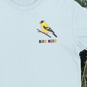 Könnte beinhalten: Ein hellblaues T-Shirt mit einer kleinen Grafik auf der linken Brustseite. Die Grafik zeigt einen gelb-schwarzen Distelfinken auf einem braunen Ast. Unter dem Vogel ist der Text BIRD NERD in bunten, einzelnen Blockbuchstaben gedruckt.