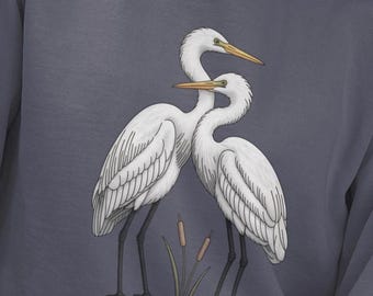 May include: A slate blue sweatshirt with a detailed illustration of two white egrets. The birds have long black legs and yellow beaks. Cattails are in the foreground. The design is centered on the front of the garment.