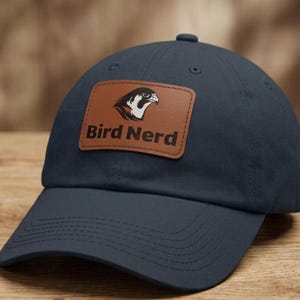 May include: A dark navy blue baseball cap featuring a rectangular brown leather patch on the front. The patch displays a black and white graphic of a bird head above the text "Bird Nerd" printed in a bold, dark brown sans-serif font.