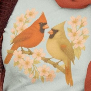 Könnte beinhalten: Hellblaues Sweatshirt mit einem Design von zwei Kardinälen auf einem Ast mit rosa Blüten. Ein Vogel ist orange, der andere gelb-grün, beide mit schwarzen Akzenten.