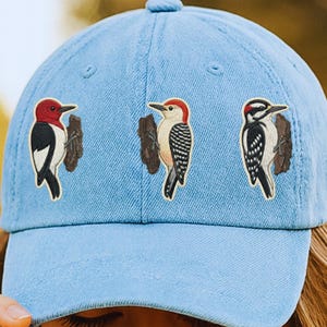 May include: Light blue baseball cap featuring embroidered woodpecker designs. The cap has three different woodpecker species, each perched on a small piece of wood. The woodpeckers have red, black, and white coloring.