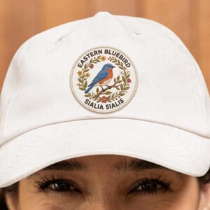 May include: A white baseball cap featuring an embroidered patch with an Eastern Bluebird design. The patch includes the text "Eastern Bluebird" and "Sialia Sialis" surrounding a blue and orange bird perched on a branch.