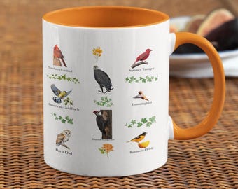 May include: White ceramic mug with an orange interior and handle. The mug features illustrations of various birds, including a Northern Cardinal, American Goldfinch, and Barn Owl, with their names printed below. The mug is a perfect gift for bird lovers.