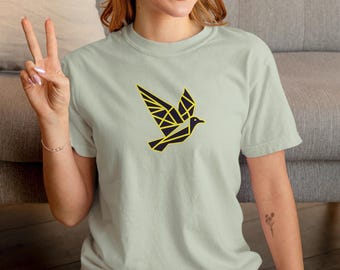 Puede incluir: Una camiseta de manga corta en color verde salvia claro con un gráfico geométrico de un pájaro en el pecho. El diseño del pájaro está compuesto por líneas azul marino oscuro y amarillo brillante, creando una representación estilizada y angular.