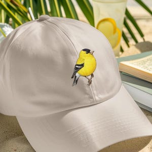 Puede incluir: Gorra de béisbol beige con una ilustración detallada de un pájaro amarillo con detalles en negro y blanco. El pájaro está posado en una rama. La gorra tiene una visera curva.