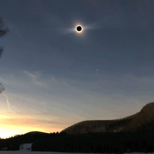 May include: A total solar eclipse with the sun's corona visible as a bright ring around the dark silhouette of the moon. The eclipse is taking place over a snowy landscape with a forest in the background.