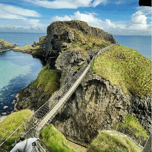 May include: A rope bridge stretches across a narrow gap between two rocky cliffs, with a person walking across the bridge. The cliffs are covered in green grass and the water below is a vibrant blue.