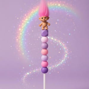 May include: A whimsical lollipop featuring a classic Troll doll with pink hair atop a stack of colorful spheres and a purple cube. The lollipop is set against a lavender background with a rainbow and glitter.