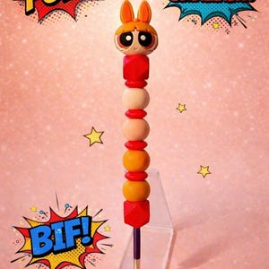 May include: A handmade pen with a cartoon character head and a series of red, orange, and beige beads. The pen is displayed on a clear stand against a pink background with comic book style text bubbles that say "POW!", "SHAZAM!", and "BIF!"
