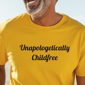May include: A yellow t-shirt with the text "Unapologetically Childfree" in a black script font. The shirt is being worn by a person wearing sunglasses, with a beach and ocean in the background.