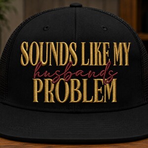 May include: A black trucker hat featuring embroidered text on the front panel. The words SOUNDS LIKE MY are stitched in gold, the word husbands is in cursive maroon thread, and the word PROBLEM is stitched in gold below the maroon text.