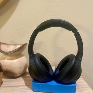 May include: Black over-ear headphones with a black headband resting on a blue stand. The headphones are on a wooden surface.