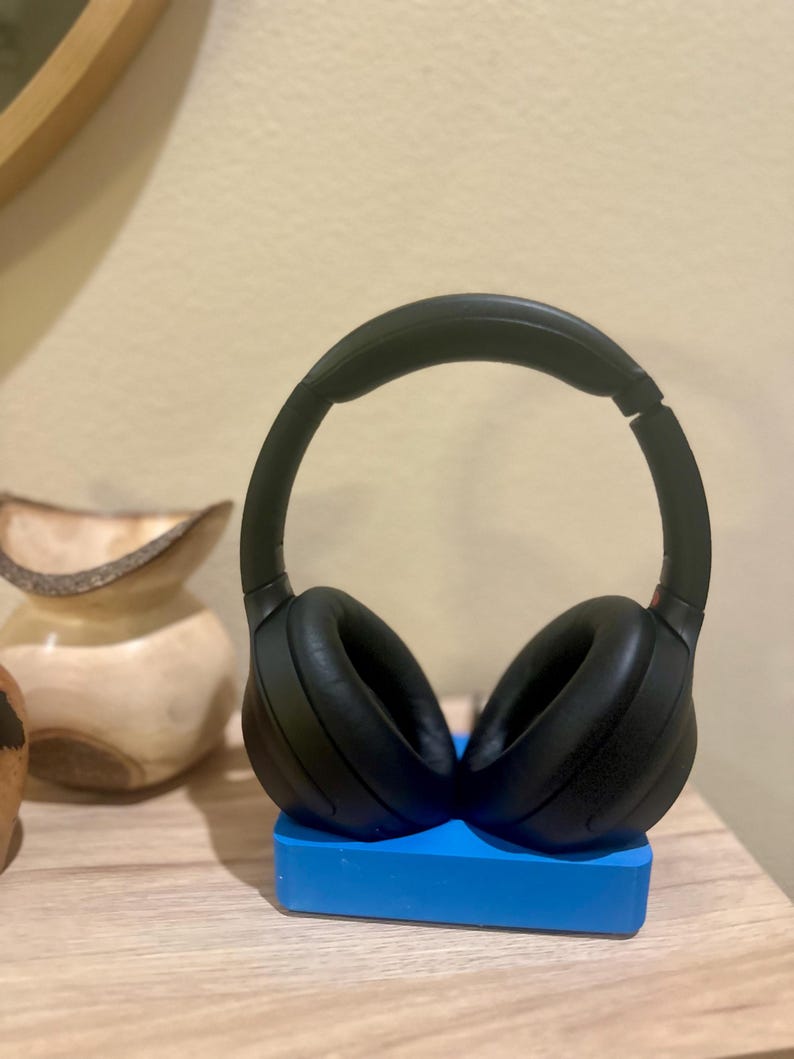 May include: Black over-ear headphones with a black headband resting on a blue stand. The headphones are on a wooden surface.
