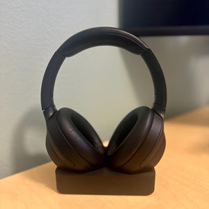 May include: Black wireless headphones with a charging stand. The headphones are on the stand and the stand is on a light wood surface.