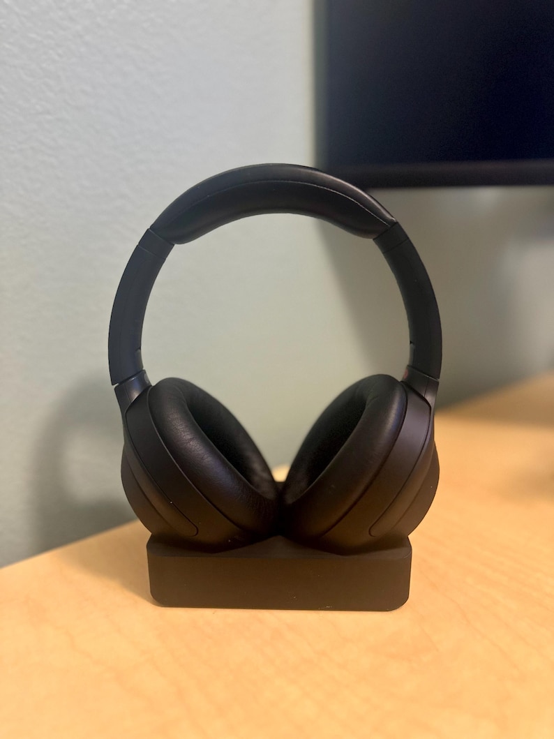 May include: Black wireless headphones with a charging stand. The headphones are on the stand and the stand is on a light wood surface.