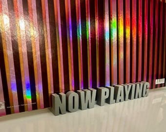 May include: Gray 3D letters spelling "NOW PLAYING" on a background of vertical iridescent stripes.