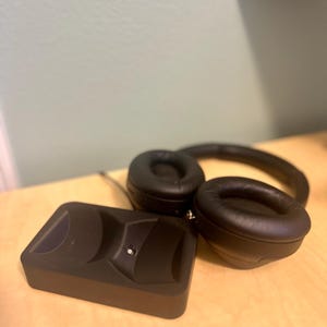 May include: Black wireless headphones with a charging base. The headphones have a black headband and earcups with a soft, padded material. The charging base is a small, rectangular device with a black finish.