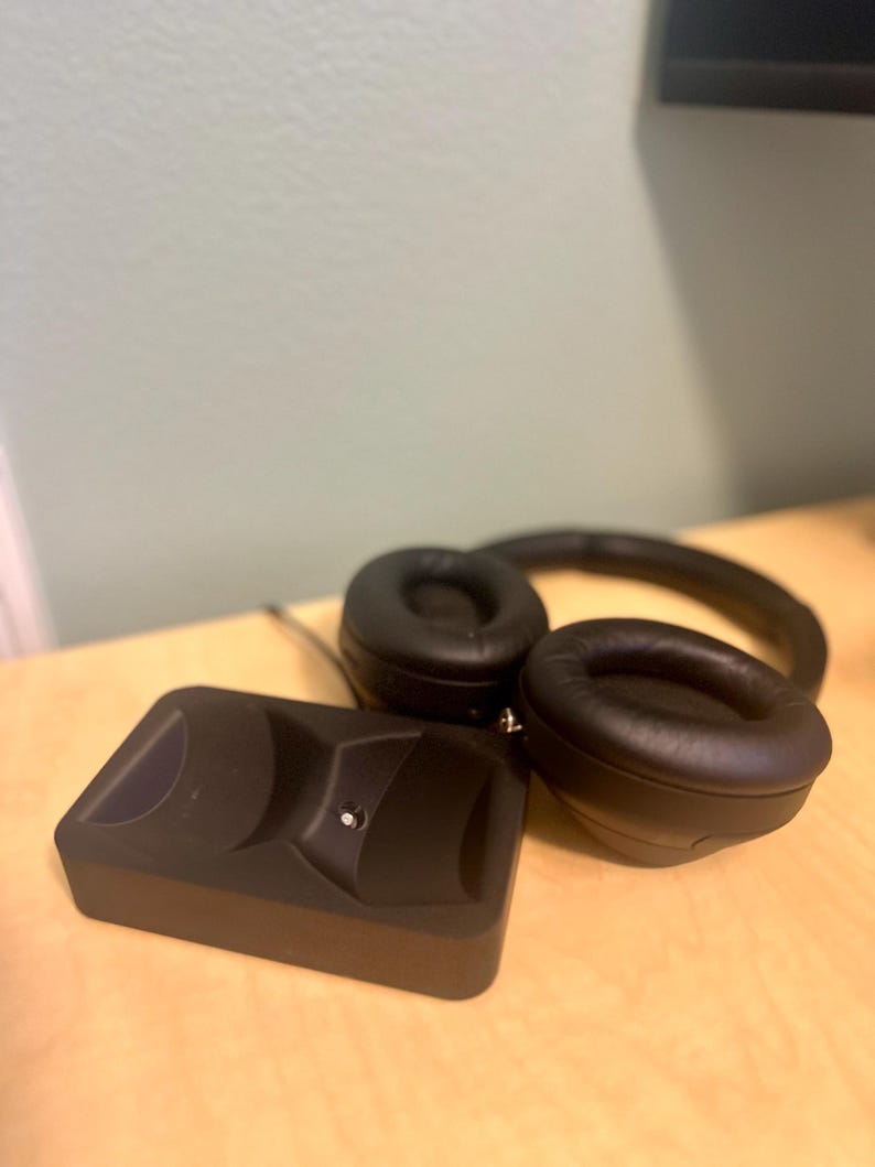 May include: Black wireless headphones with a charging base. The headphones have a black headband and earcups with a soft, padded material. The charging base is a small, rectangular device with a black finish.