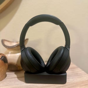 May include: Black wireless headphones with a charging base. The headphones are on the base and the earcups are facing outwards.