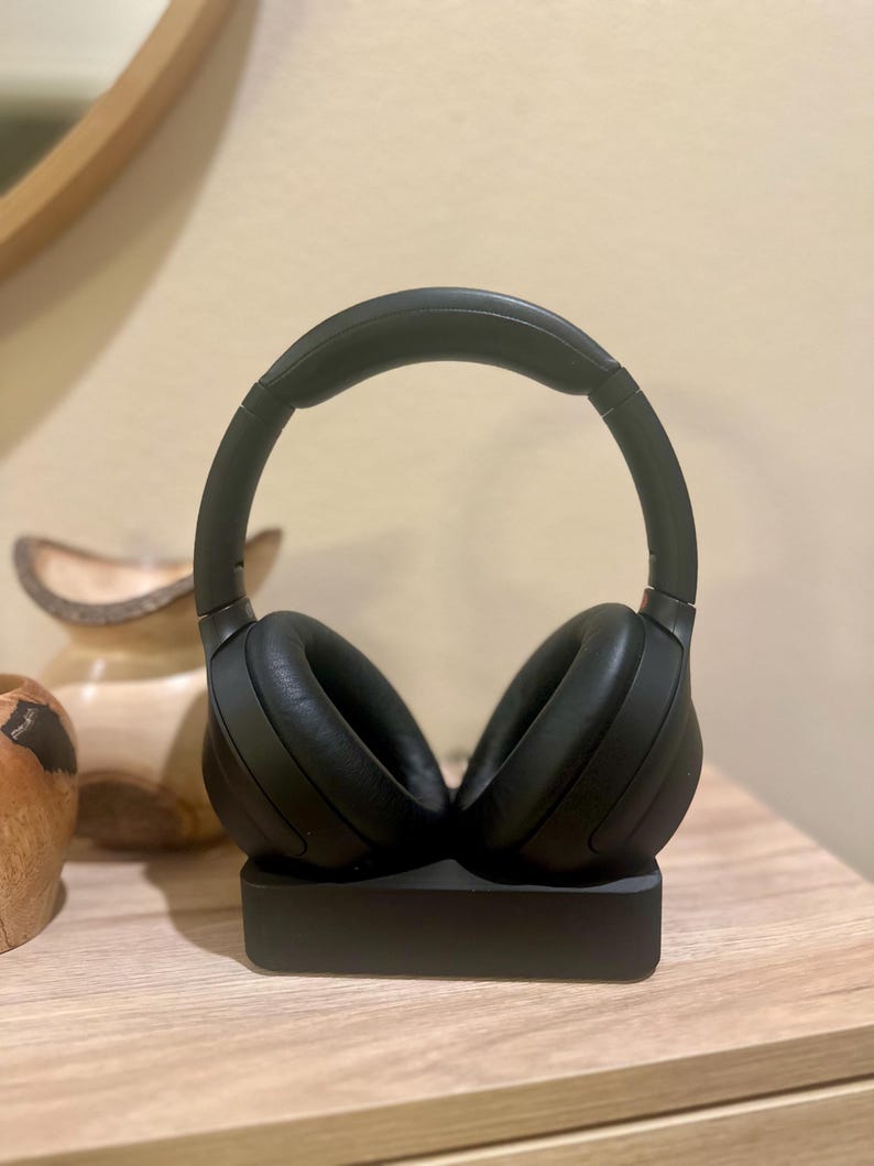 May include: Black wireless headphones with a charging base. The headphones are on the base and the earcups are facing outwards.