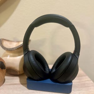 May include: Black over-ear headphones with a padded headband resting on a blue stand. The headphones are on a wooden surface.