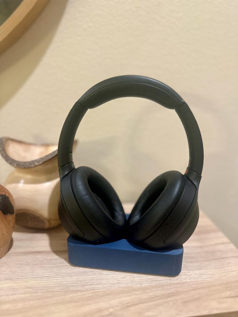 May include: Black over-ear headphones with a padded headband resting on a blue stand. The headphones are on a wooden surface.