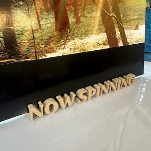 May include: A close-up of a record player stand made of wood with the words "NOW SPINNING" in raised letters.