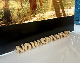 May include: A close-up of a record player stand made of wood with the words "NOW SPINNING" in raised letters.
