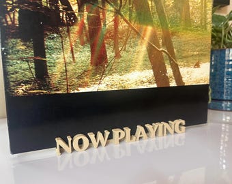Pode incluir: Uma placa de madeira com as palavras "NOW PLAYING" em letras maiúsculas está em frente a uma imagem emoldurada de uma cena de floresta. A imagem tem uma borda preta e o reflexo da placa é visível na superfície abaixo.