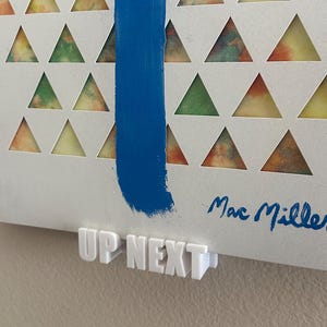May include: A white vinyl record with a blue stripe down the centre. The record has a pattern of colourful triangles cut out of the vinyl. The record is titled "Up Next" and is signed "Mac Miller".