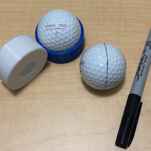 May include: A white roll of tape, a black permanent marker, and two white golf balls. One golf ball has the text "KIRKLAND Signature 2" printed on it. The other golf ball has a black line drawn across it.