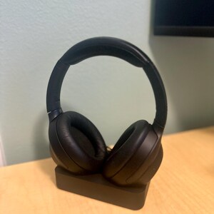 May include: Black wireless headphones with a charging stand. The headphones are on the stand with the earcups facing out.