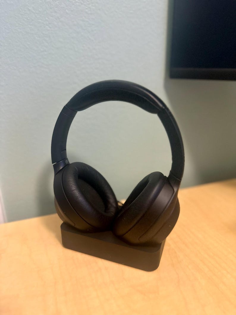 May include: Black wireless headphones with a charging stand. The headphones are on the stand with the earcups facing out.