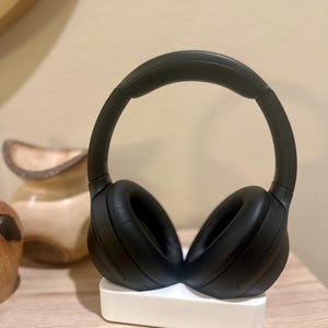 May include: Black wireless headphones with a white charging base. The headphones are on the base with the earcups facing out.