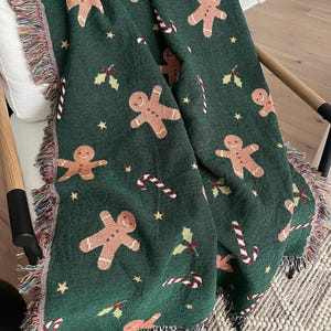 Cozy green woven Christmas throw blanket with gingerbread men, candy canes, and holly draped over a chair — festive holiday décor and winter gift idea.