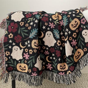 May include: A black Halloween-themed throw blanket with a repeating pattern of smiling ghosts, jack-o'-lanterns, flowers, stars, and moons. The blanket has a fringed edge with a mix of colors.