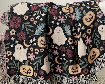 Puede incluir: Una manta negra con temática de Halloween con un patrón repetido de fantasmas sonrientes, calabazas, flores, estrellas y lunas. La manta tiene un borde con flecos.