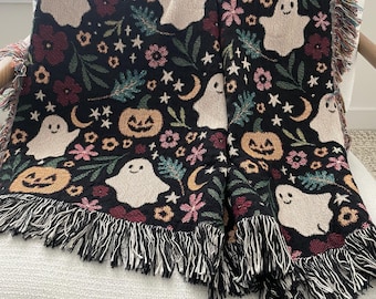Puede incluir: Una manta tejida con fondo negro y un patrón repetitivo de fantasmas beige, calabazas naranjas, flores rosas y rojas, hojas verdes, lunas crecientes amarillas y pequeñas estrellas blancas, terminada con un borde de flecos.