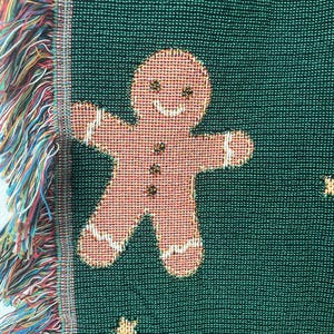 Close-up of green woven Christmas blanket featuring a smiling gingerbread man with gold stars and soft multicolored fringe edge — festive holiday textile detail.