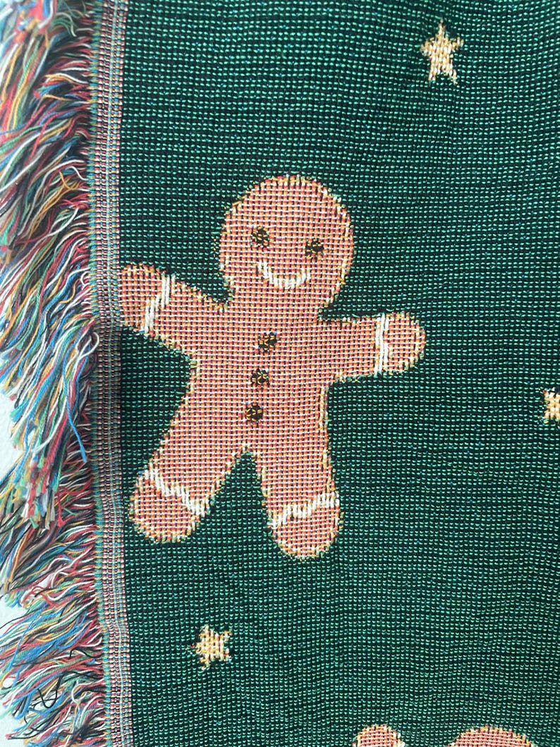 Close-up of green woven Christmas blanket featuring a smiling gingerbread man with gold stars and soft multicolored fringe edge — festive holiday textile detail.