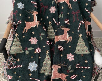 Puede incluir: Una manta tejida de color verde oscuro con un patrón repetitivo de renos de color marrón claro, árboles de Navidad verdes, lazos rojos, bastones de caramelo y adornos en forma de estrella. La manta tiene un borde con flecos multicolores.