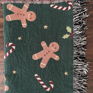Green woven Christmas throw blanket with gingerbread men, candy canes, and holly, featuring soft black-and-white and multicolored fringe edges for a festive touch.