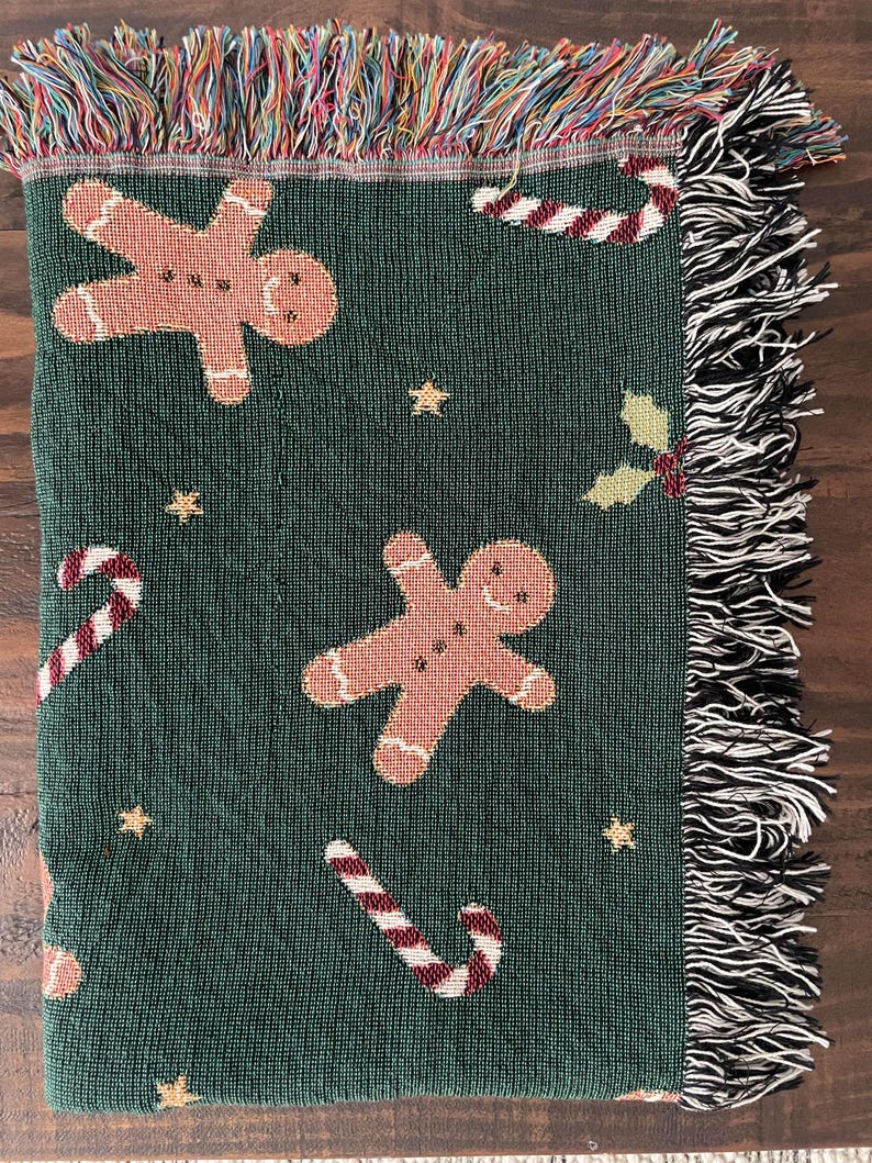 Green woven Christmas throw blanket with gingerbread men, candy canes, and holly, featuring soft black-and-white and multicolored fringe edges for a festive touch.