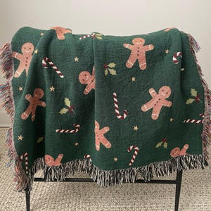 Green woven Christmas throw blanket with gingerbread men, candy canes, holly, and stars draped over a chair — festive holiday décor and cozy winter gift.