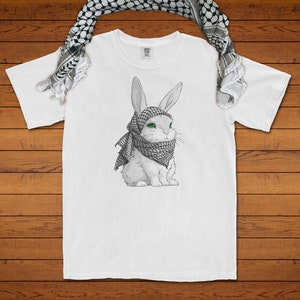 May include: A white t-shirt featuring a graphic illustration of a white rabbit with bright green eyes wearing a black and white chequered keffiyeh scarf. A matching black and white chequered keffiyeh scarf is draped over the top of the t-shirt.