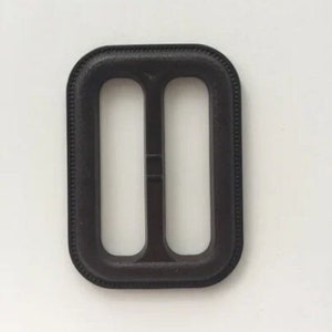 Puede incluir: Una hebilla de plástico negra con dos ranuras para una correa. La hebilla tiene una superficie lisa y ligeramente texturizada.